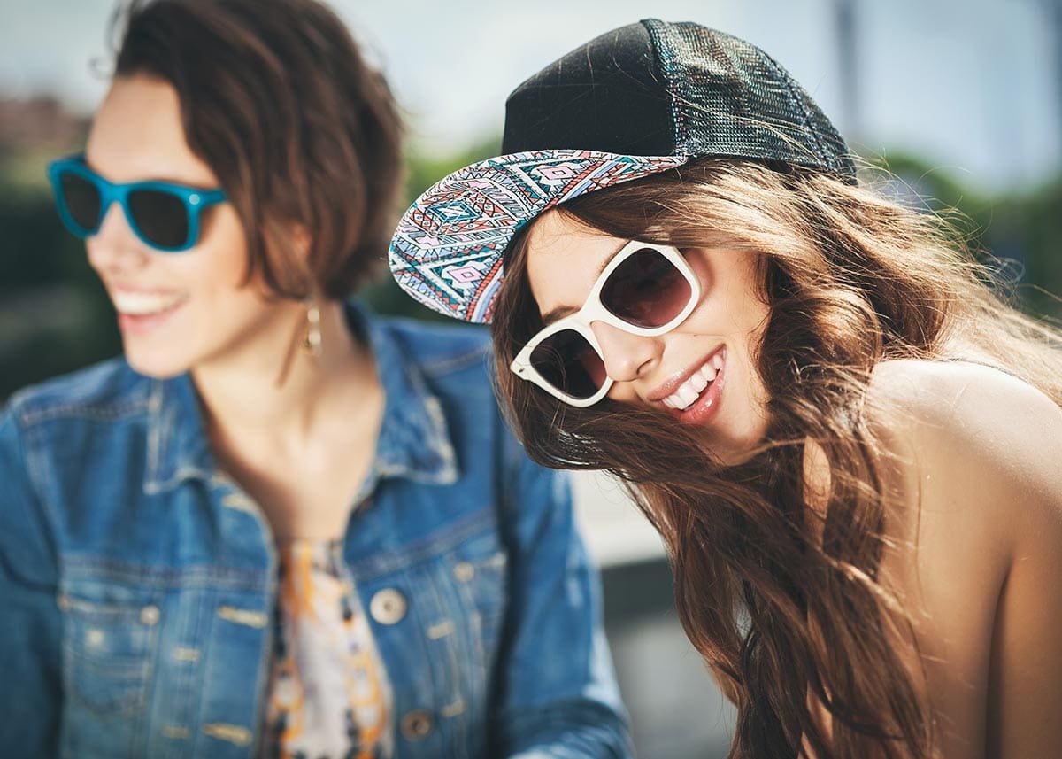 Two smiling women in sunglasses outdoors: one in a denim jacket with blue frames, one in a patterned snapback cap and white sunglasses.