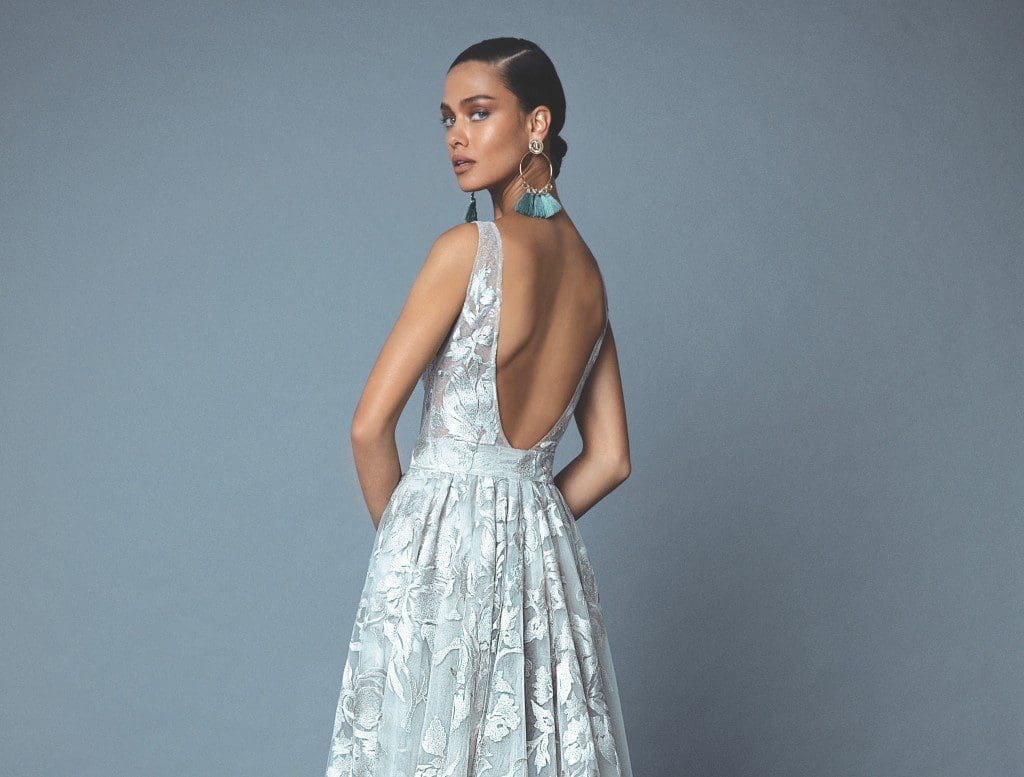 A woman in a pale mint floral lace gown with an open back and turquoise tassel earrings, against a gray-blue backdrop.