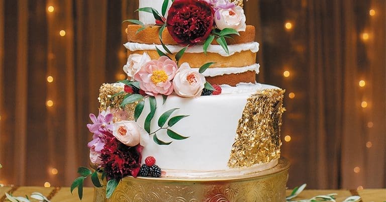 A tiered wedding cake with white fondant, gold leaf accents, and fresh peonies, roses and berries on a gold stand.