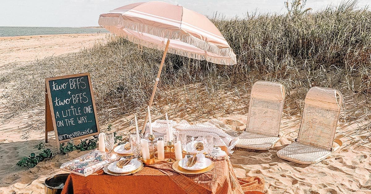 A beach picnic table set with candles, plates and a pink fringed umbrella, beside a chalkboard sign reading "two BFFs + two BFFs, a little one on the way."