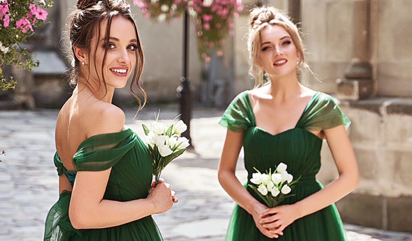 Two bridesmaids in emerald green tulle gowns hold white tulip bouquets on a cobblestone street.