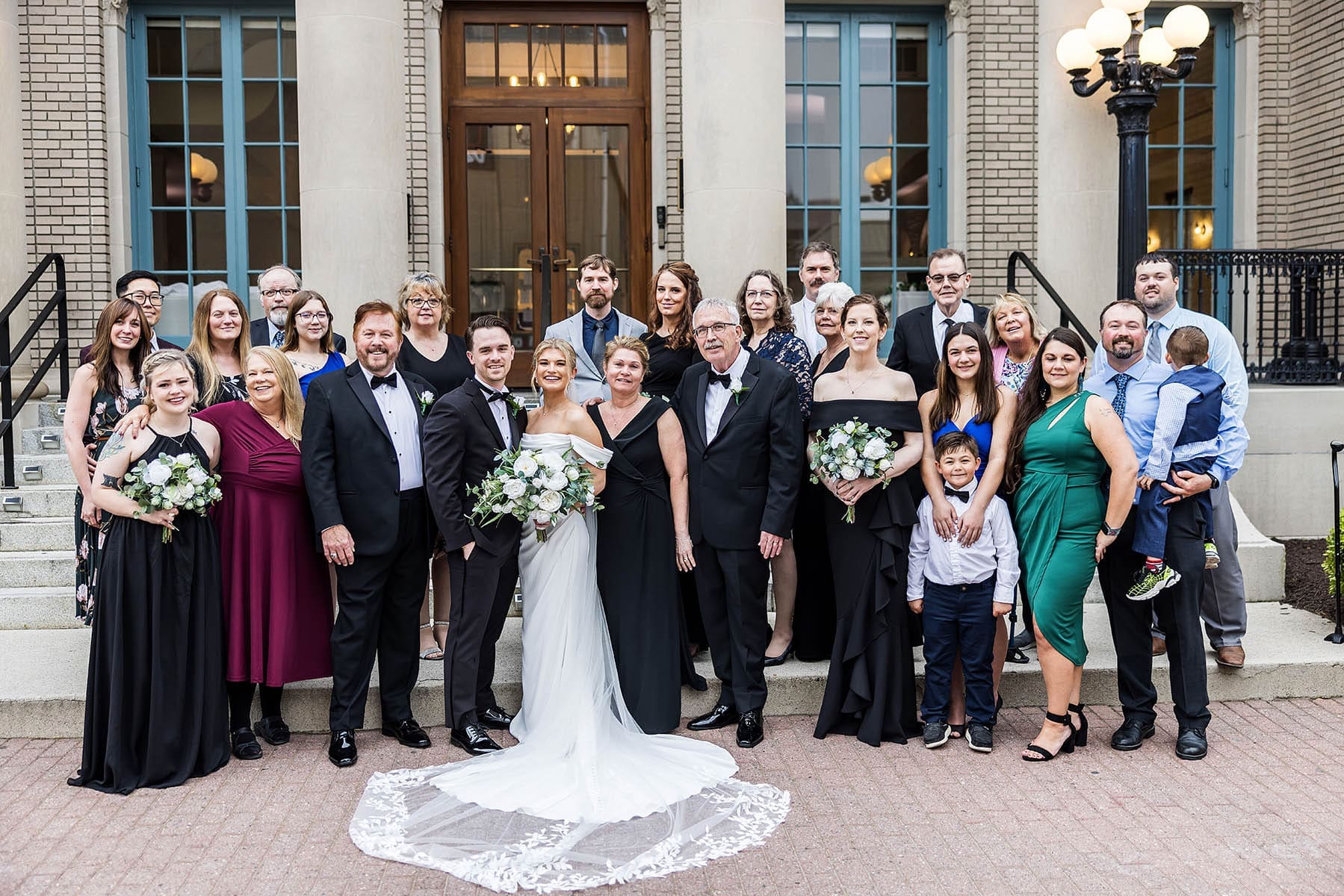 A large group of wedding guests in formal attire poses on steps outside a brick building with a bride in a lace-trimmed gown.
