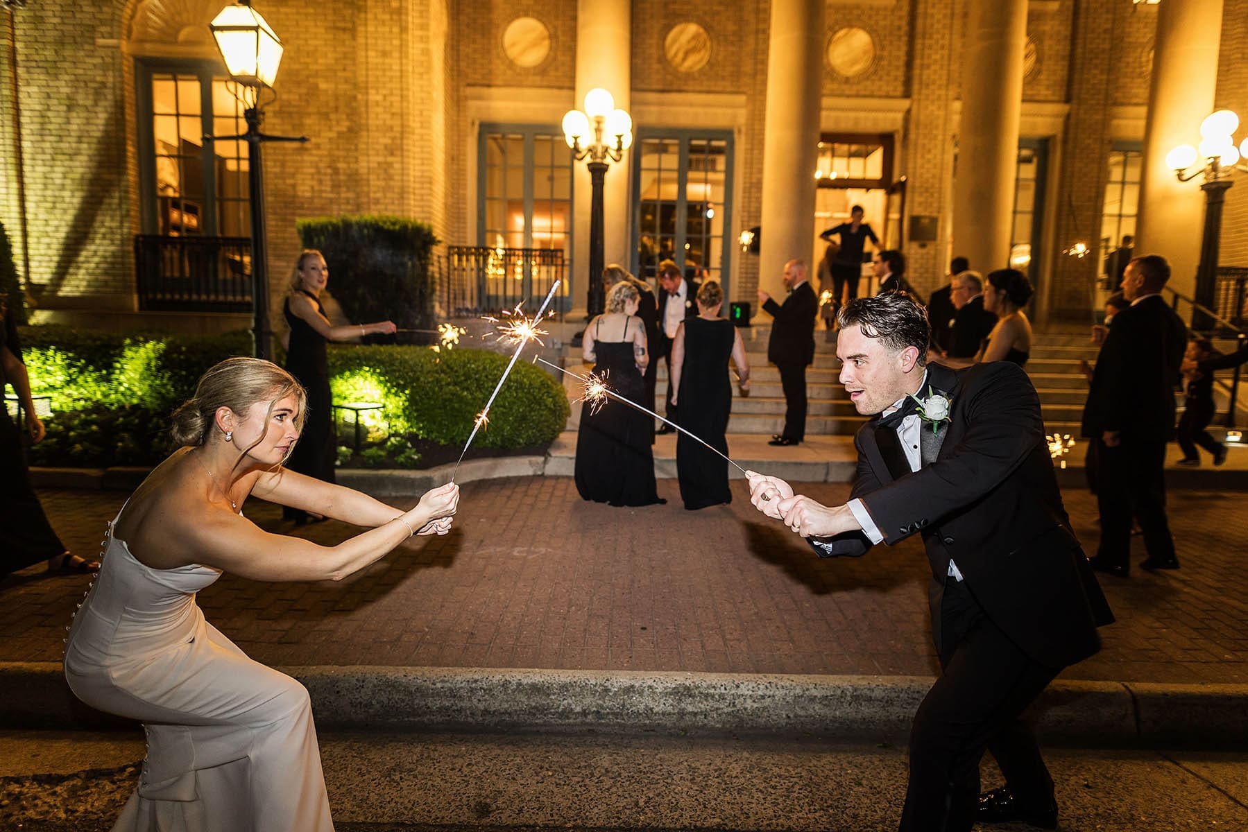 A bride and groom hold sparklers facing each other while guests in formal attire gather on a lit brick courtyard at night.