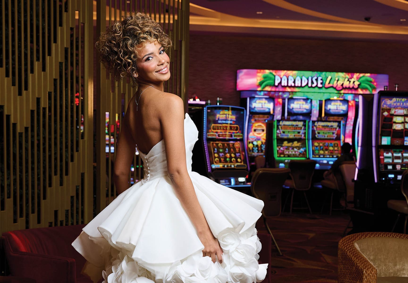 A woman with curly updo hair in a strapless white ballgown with ruffled skirt smiles over her shoulder in a casino with slot machines.