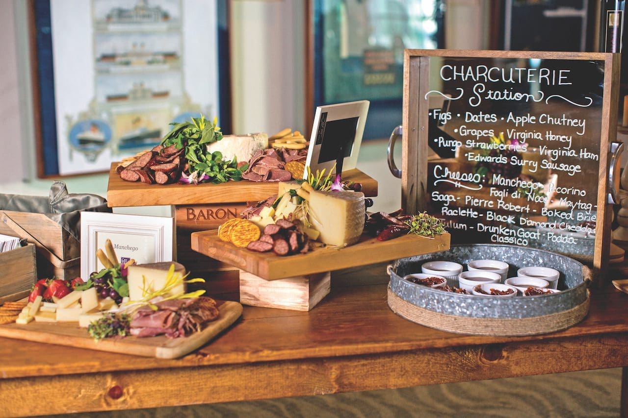 A charcuterie station with cured meats, cheeses, and fruit on wooden boards, beside a chalkboard menu reading "Charcuterie Station."