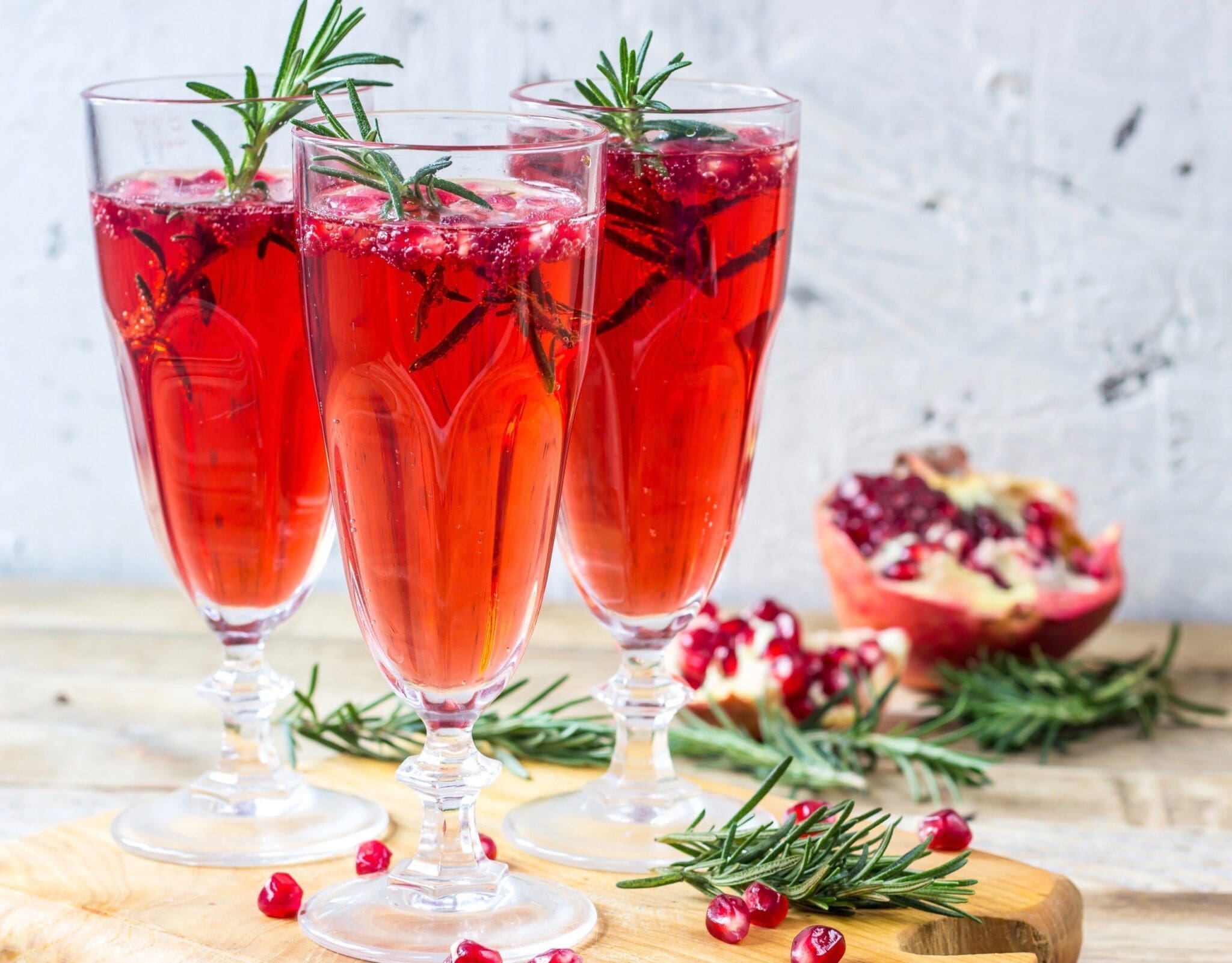 Three stemmed flutes of red pomegranate cocktail garnished with rosemary sprigs, with a halved pomegranate and pomegranate seeds nearby.