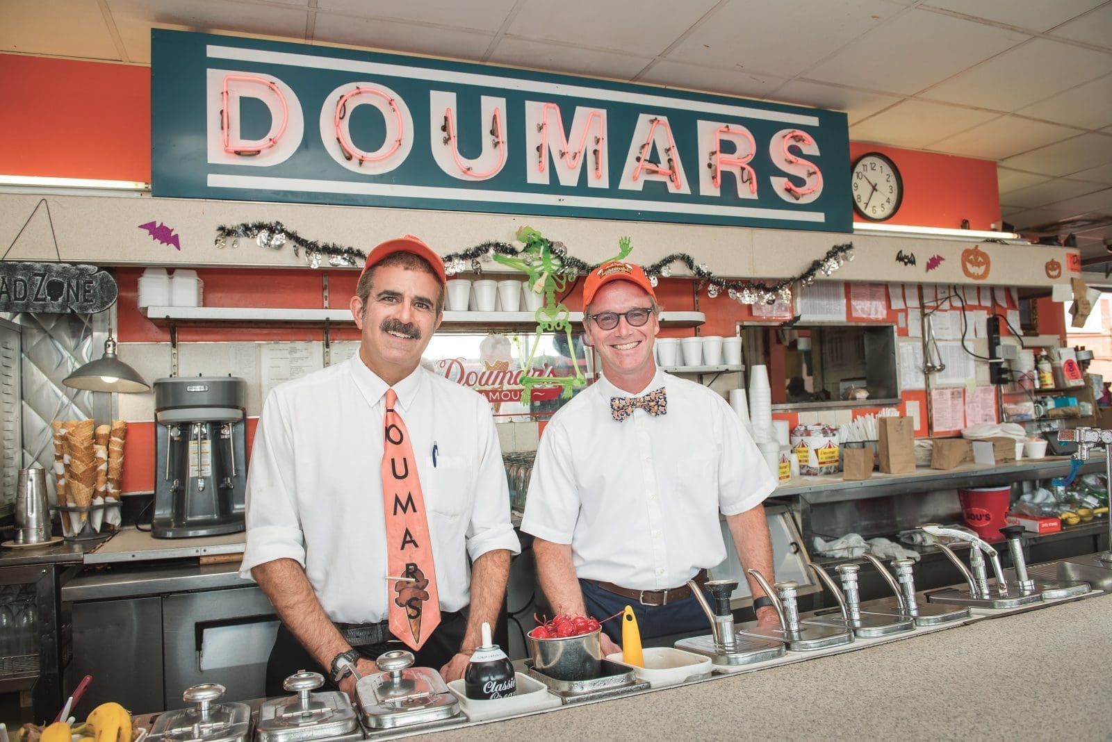 Two smiling men in white shirts and orange caps stand behind the Doumars counter beneath its neon sign.