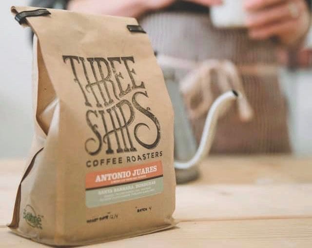A kraft paper coffee bag labeled "Three Ships Coffee Roasters" and "Antonio Juarez" sits on a wooden table near a gooseneck kettle.