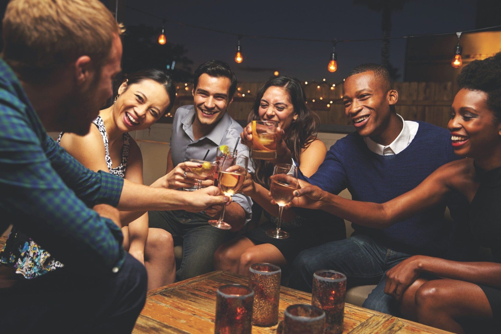 Six friends toast with cocktails at a wooden table under string lights on a rooftop at night.
